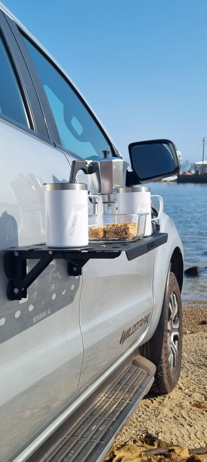 A black car-side shelf mounted on the side of a silver vehicle, holding a white insulated mug, a clear cup with a lid, and a small container of snacks, parked beside water under a blue sky.