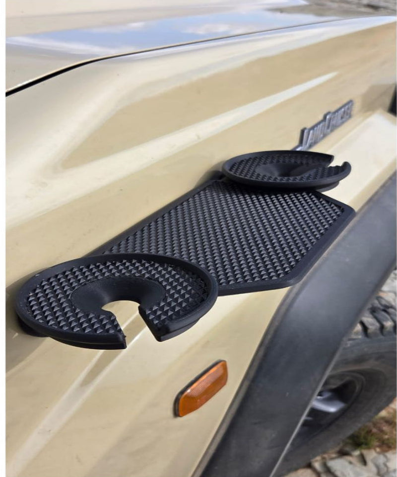 A close-up of a beige Land Cruiser’s front fender with a black textured magnetic tray attached. The tray features two circular cup holders with open slots, designed to securely hold drinks or small items while parked. The accessory has a rugged, non-slip surface and fits neatly against the vehicle’s body, highlighting its practical outdoor and overlanding use.
