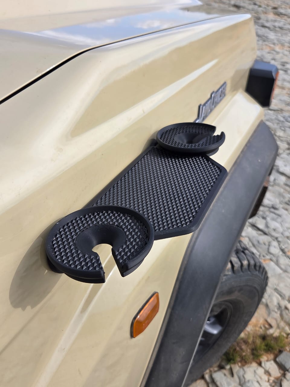A close-up of a beige Land Cruiser’s front fender with a black textured magnetic tray attached. The tray features two circular cup holders with open slots, designed to securely hold drinks or small items while parked. The accessory has a rugged, non-slip surface and fits neatly against the vehicle’s body, highlighting its practical outdoor and overlanding use.
